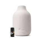 A white ribbed ceramic diffuser bottle with a small dark amber essential oil bottle placed in front, shown on a clean white background. The diffuser has the PURAS logo printed on the front.