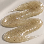 Close-up of a glossy beige body scrub swatch with fine brown exfoliating particles, spread in a curved pattern on a cream background.
