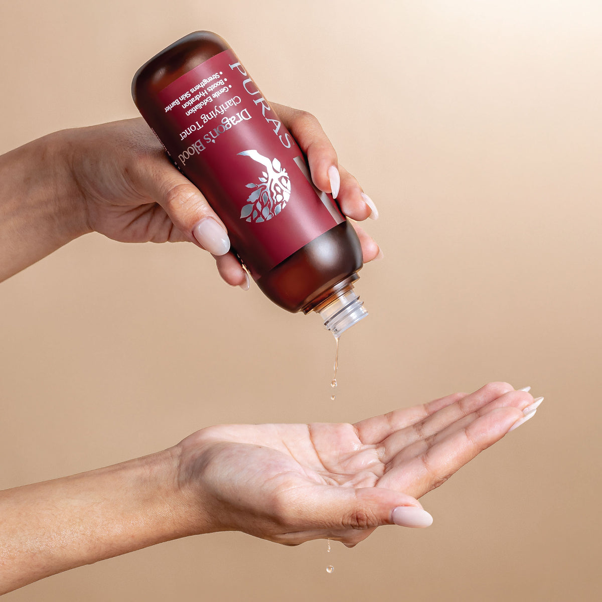 Hand holding a Puras Dragon's Blood Clarifying Toner with a label, pouring liquid into another hand against a beige background