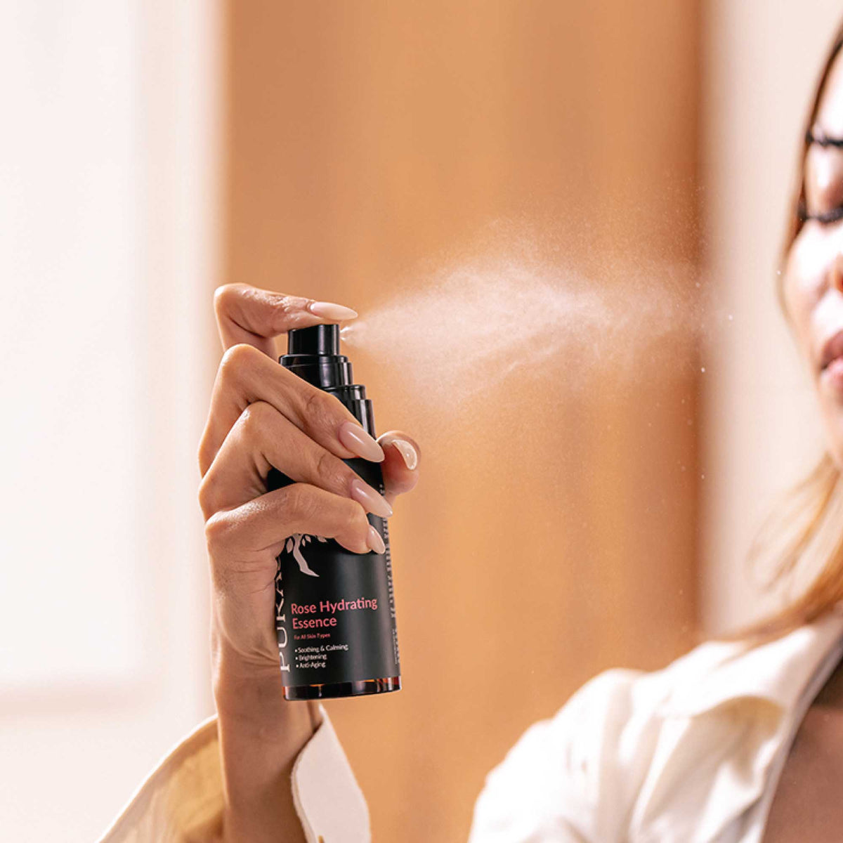 Person holding a bottle of 'Rose Hydrating Essence' and spraying it.
